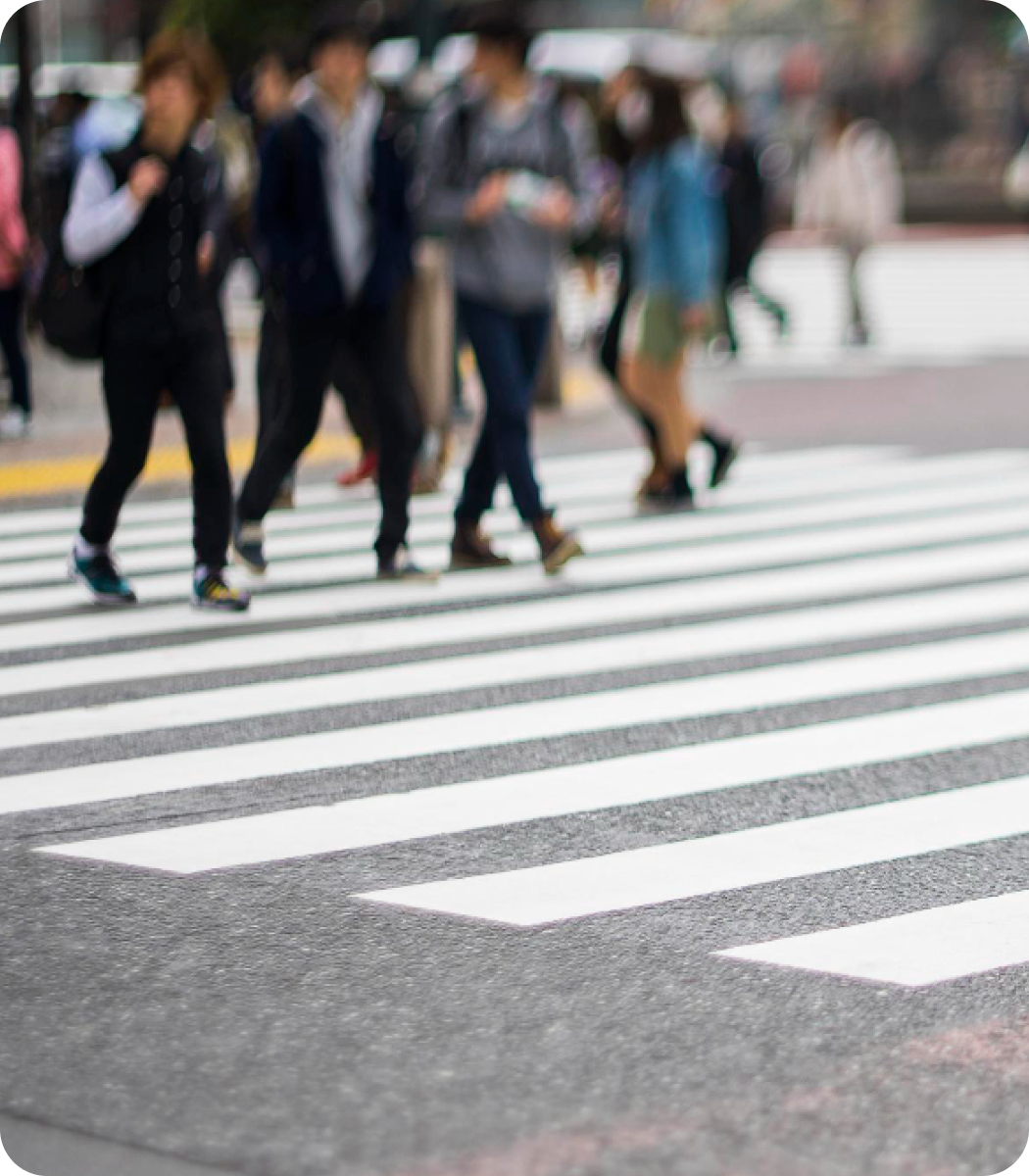 Close-up horizontal dari penyeberangan zebra dengan garis-garis putih tebal di atas aspal abu-abu gelap. Gambar pejalan kaki yang sedikit buram terlihat berjalan di latar belakang.