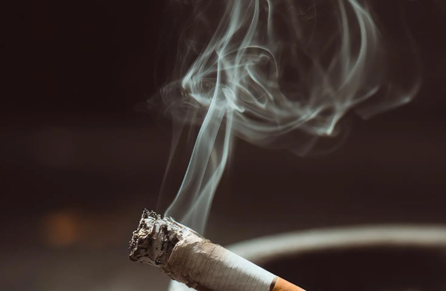 Jarak dekat rokok yang menyala di asbak engan ujungnya berada di tepi asbak dengan gumpalan asap tebal melengkung ke atas di latar belakang yang gelap. Ujung rokok tertutup abu dan bara.