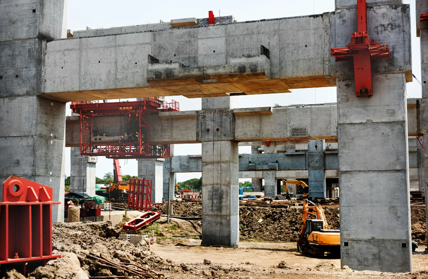 Area konstruksi yang sibuk menunjukkan struktur beton pracetak besar yang belum selesai, kemungkinan untuk jalan layang atau jembatan, dengan tanah galian yang luas, material, dan alat berat oranye yang terlihat di bawah. Sumber kontruksi ini dapat menimbulkan polusi Partikulat Materi 10 (PM10).