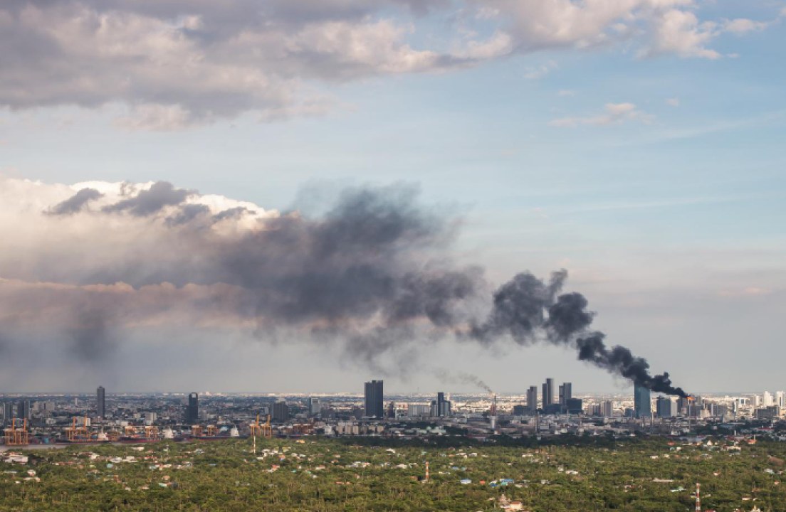 Pemandangan udara yang luas (bird's-eye view) dari kota yang padat dengan gedung-gedung pencakar langit dan area hijau yang luas di latar depan. Asap hitam pekat mengepul dari salah satu gedung tinggi di cakrawala yang diduga adalah gedung pabrik, membentuk gumpalan besar yang menyebar di bawah langit berawan. Asap ini dapat menimbulkan zat polusi Nitrogen Oksida.