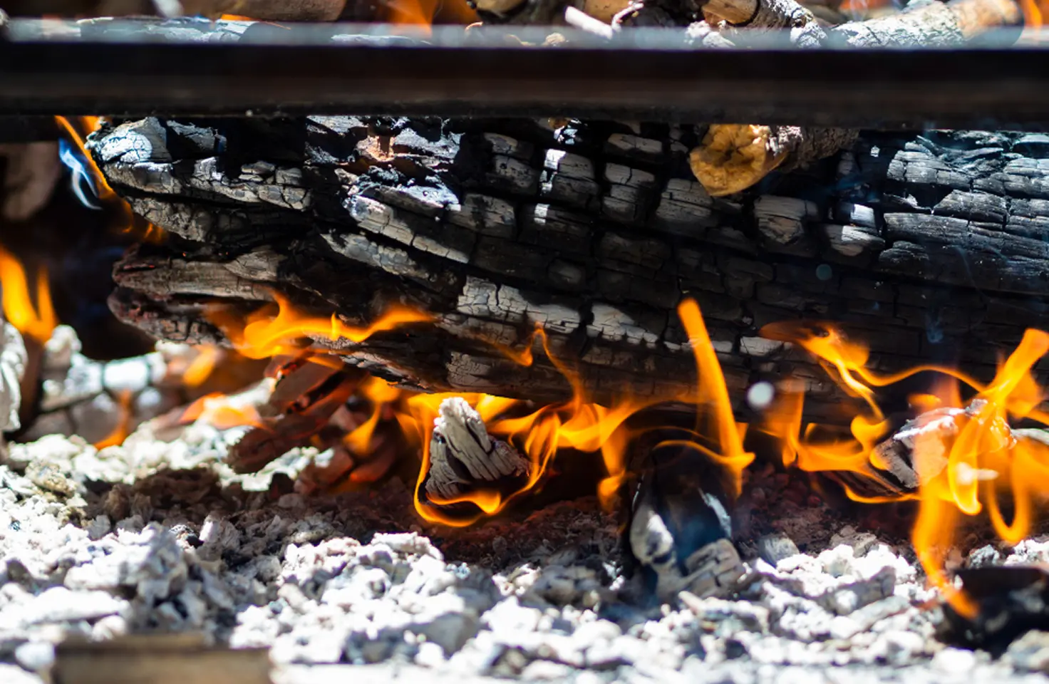 Gambar dekat kayu yang terbakar di atas api unggun atau panggangan dengan bara api dan abu putih di bawah. Kayu yang terbakar memiliki tekstur hangus, dan nyala api oranye terang mengelilinginya. Hasil pembakaran ini dapat menimbulkan zat polusi Sulfir Dioksida.